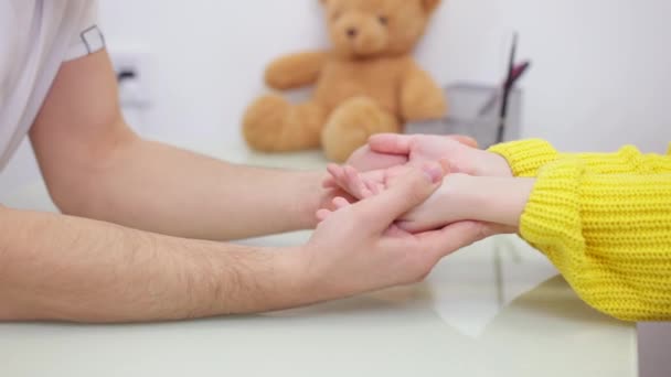 Close-up of male hands holding child palms with blurred teddy bear at background. Unrecognizable Middle Eastern doctor calming down girl on appointment. Pediatric care and medicine. — Stock Video
