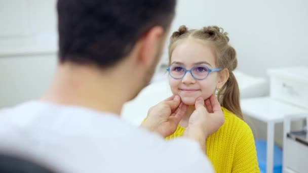 Shooting over doctor shoulder of cute smiling girl looking at pediatrician checking thyroid. Positive Caucasian patient undergoes medical examination in hospital. Health care and childhood. — Stock Video