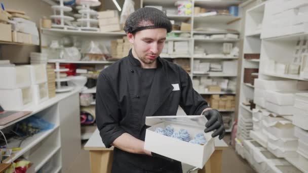 Positiv gut aussehender kaukasischer junger Mann posiert mit gekochtem leckerem Dessert im Süßwarenladen. Porträt eines Bäckers in der Mitte, der lächelnd in die Kamera blickt und eine Pappschachtel mit leckeren Muffins hält. — Stockvideo