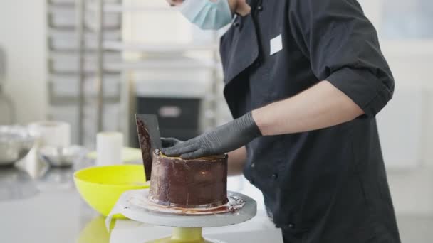 Seitenansicht eines nicht wiederzuerkennenden Mannes mit Coronavirus-Gesichtsmaske, der Schokolade auf Kuchen auf einem Drehtisch verteilt. Konzentrierte männliche Konditormeister dekorieren Bäckerei in Küche drinnen. — Stockvideo