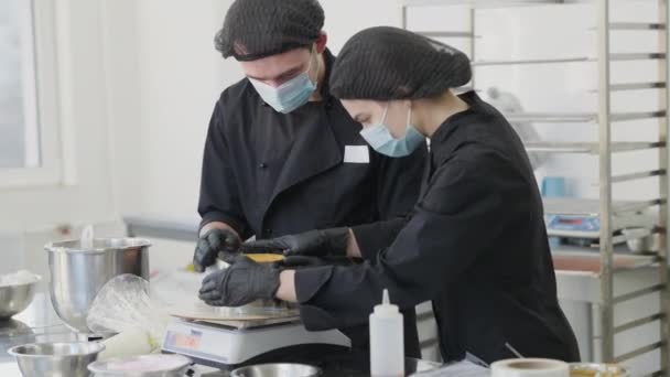 Portret białego mężczyzny pomagającego kobiecie gotować ciasto w kuchni handlowej. Profesjonalny cukiernik uczy stażystę w cukierni. Pieczenie i kuchnia na pandemię koronawirusa. — Wideo stockowe