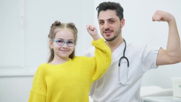 O fată caucaziană fericită în ochelari de vedere și un bărbat pozitiv din Orientul Mijlociu care arată un gest de forță privind camera foto. Portret de medic vesel și pacient pozând în spital în interior. Conceptul de sănătate . — Videoclip de stoc