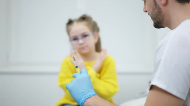 Professionele kinderarts uit het Midden-Oosten met Covid-19 vaccin in een injectiespuit die praat met wazig bang blank meisje dat op de achtergrond zit met gekruiste handen. Bang patiënt in ziekenhuis afdeling binnenshuis — Stockvideo