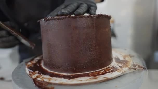 Mains blanches masculines en gants noirs tournant table tournante ajoutant garniture de chocolat sur délicieux gâteau cuit au four. Confiseur boulanger méconnaissable décorant de délicieuses pâtisseries dans la cuisine commerciale. — Video