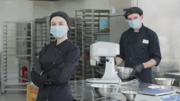 Retrato de una joven y alegre mujer delgada en máscara facial Covid-19 y uniforme de cocinero mirando a la cámara sonriendo con el hombre borroso posando al fondo. Chef en cocina comercial en tienda de dulces en pandemia. — Vídeos de Stock