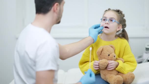 Pédiatre Moyen-Orient joyeux distrayant fille caucasienne mignonne prenant test Covid. Médecin positif apaisant petit patient à l'examen médical à l'hôpital sur la pandémie de coronavirus. — Video
