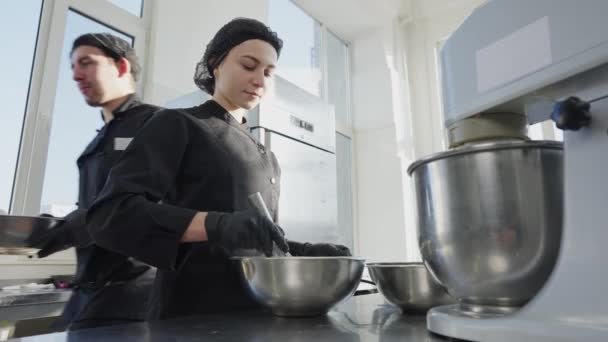 Młoda zmęczona kobieta mieszająca ciasto, wycierająca czoło i patrząca na uśmiechniętą kamerę. Człowiek przechodzący w tle ze składnikami. Portret pozytywnego pięknego cukiernika w kuchni sklepu cukierniczego. — Wideo stockowe