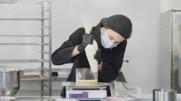 Portrait of focused female confectioner adding layer on baked cake dough standing on kitchen scales. Young slim Caucasian woman in coronavirus face mask cooking dessert indoors. — Stock Video