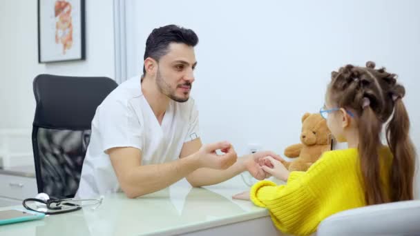 Plan médian d'un pédiatre masculin joyeux tenant la main d'un patient parlant et souriant. Positif beau médecin du Moyen-Orient bavarder avec fille caucasienne sur rendez-vous. Consultation et médecine. — Video