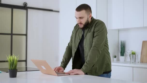 Portrait of thoughtful concentrated Caucasian man standing in kitchen with laptop. Bearded young inspired guy thinking of ideas at home indoors. New normal on pandemic lockdown. — Vídeo de Stock