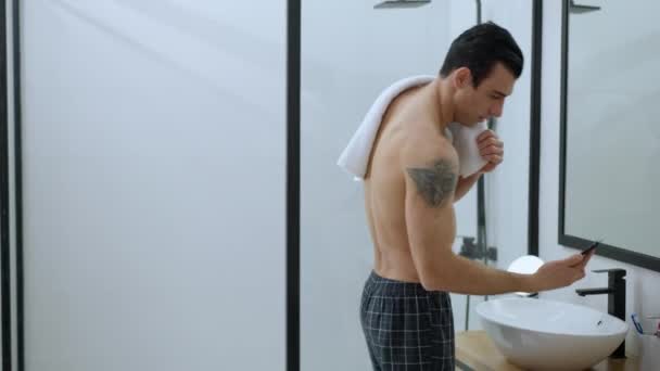 Portrait of happy handsome man receiving message in bathroom in the morning. Positive tattooed Middle Eastern guy messaging on smartphone at home indoors. Device addiction concept. — Vídeo de Stock
