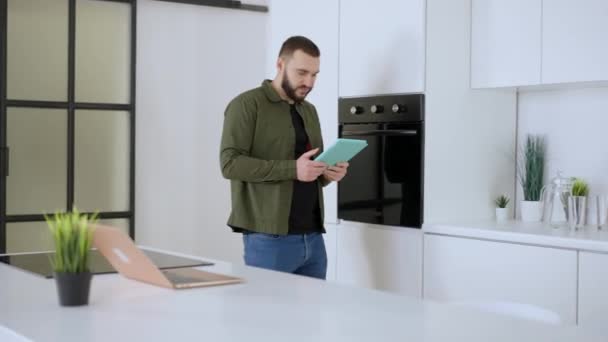 Hombre absorto confiado entrando en la cocina sosteniendo tableta deslizando las redes sociales. Retrato de un caucásico guapo concentrado navegando por Internet en casa. Dispositivo adicción estilo de vida. — Vídeos de Stock