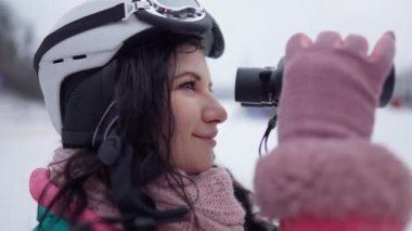 Kayak kasklı güzel bir kadının dürbünle bakışlarının yan görüntüsü. Kendine güveni tam güzel beyaz kayakçının yakından çekilmiş yüzü. Açık havada kışı seyrediyor..