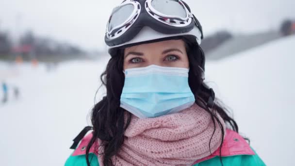 Retrato de cerca de la joven mujer caucásica con máscara facial coronavirus y traje de esquí mirando a la cámara posando al aire libre. Cabeza de turista mujer descansando en el complejo invernal nevado en la pandemia de Covid-19. — Vídeos de Stock