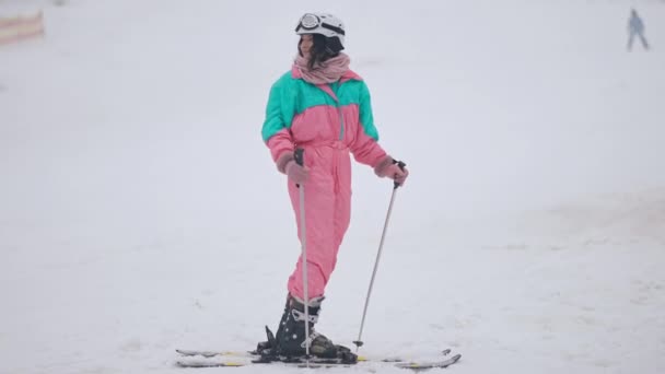Szerokie ujęcie szczęśliwej szczupłej kobiety w kombinezonie narciarskim machającej i odjeżdżającej na śnieżnych stokach. Portret wesołego białego narciarza korzystającego z wypoczynku zimowego w kurorcie na świeżym powietrzu. — Wideo stockowe