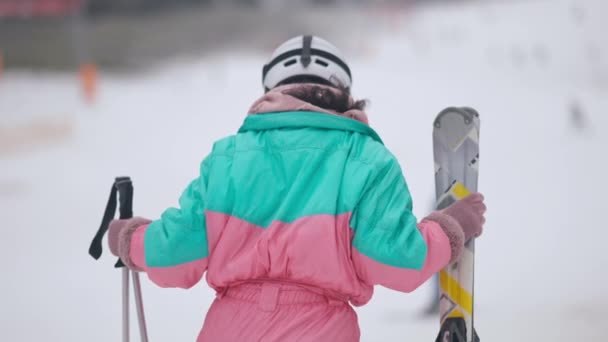 Visão traseira da mulher magra em terno de esqui e capacete andando em câmera lenta com esquis e bastões de esqui. Relaxado jovem esquiador feminino em encostas nevadas ao ar livre. Turismo e lazer de inverno. — Vídeo de Stock