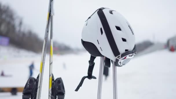 Capacete de esqui em postes com esquis e resort de inverno borrado no fundo. Equipamento de esqui ao ar livre. — Vídeo de Stock