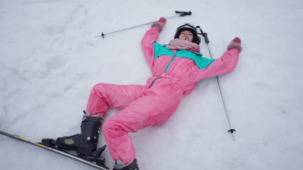 Szeroki widok na zrelaksowaną białą kobietę w kombinezonie narciarskim leżącą na śniegu na świeżym powietrzu. Szczęśliwy beztroski szczupły narciarz turystyczny odpoczywający w ośrodku zimowym. Wypoczynek i styl życia. — Wideo stockowe