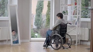 Aynaya bakıp tekerlekli sandalyeyle uzaklaşan üzgün, genç, beyaz adamın geniş bir görüntüsü. Çaresiz, felçli, felçli bir adam evden dışarı çıkıyor. Engelli ve engelli kavramı.