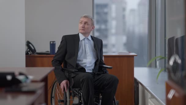 Portrait of thoughtful senior grey-haired man in wheelchair indoors. Sad disabled Caucasian male employee at workplace in office. Inclusive business and lifestyle concept. — Stock Video