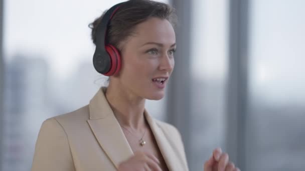 Mujer hermosa delgada positiva en auriculares disfrutando de la música bailando en la ventana. Retrato de feliz relajado empleado CEO caucásico divirtiéndose en el descanso en la oficina. Estilo de vida y aficiones. — Vídeo de stock