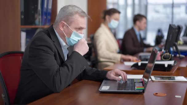 Sidovy av sjuk senior kaukasisk man i coronavirus ansikte mask hosta med suddiga medarbetare sitter vid datorer i bakgrunden. Sjuk manlig anställd på kontoret inomhus på Covid-19 pandemiutbrott. — Stockvideo