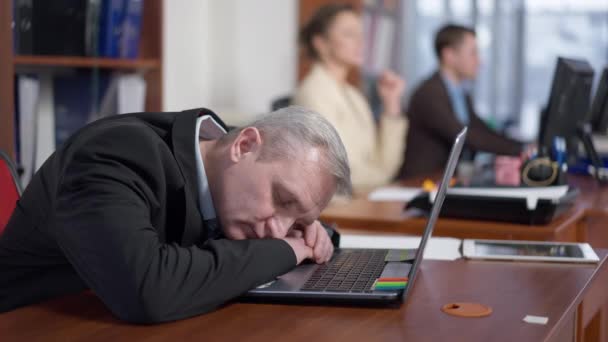 Измученный, перегруженный работой старший мужчина спит на рабочем столе с размытыми коллегами, работающими на заднем плане. Портрет уставшего кавказского работника в помещении. Переутомление и бизнес. — стоковое видео