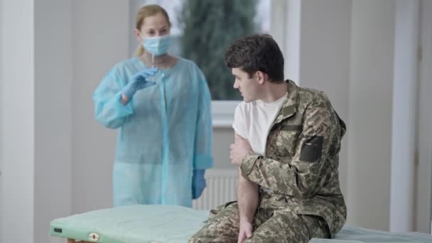 Soldado joven nervioso sentado en el sofá médico en el interior como enfermera borrosa o médico sosteniendo la jeringa con la vacuna contra el coronavirus de pie en segundo plano. Vacunación Covid-19 en el ejército. — Vídeos de Stock