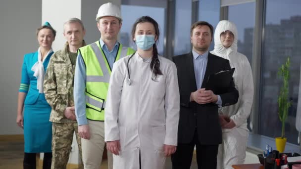Dokter wanita Kaukasia muda yang cantik atau perawat di Covid-19 topeng wajah saling bersilang tangan melihat kamera dengan orang-orang dari profesi yang berbeda berpose di latar belakang. Kedokteran dan perawatan kesehatan pada pandemi. — Stok Video