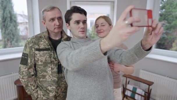 Joven positivo tomando selfie con padre militar y madre sonriente en el interior. Hijo caucásico adulto fotografiando a la familia en el teléfono inteligente en casa. Unidad y estilo de vida familiar. — Vídeo de stock