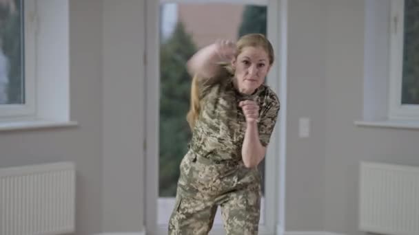 Mellanslag av självsäker kvinnlig soldat i uniform boxning skugga tittar på kameran. Porträtt av koncentrerad kaukasisk medelålders kvinna som tränar hemma på kvällen. Feminism och jämställdhet. — Stockvideo