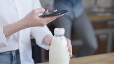 Tanımlanamayan bir kadının, akıllı telefonuyla soru cevap kodunu süt şişesinde taramasına yakın çekim. Genç, sıska bayan modern teknolojileri günlük hayatta kullanıyor. Evdeki mutfakta..