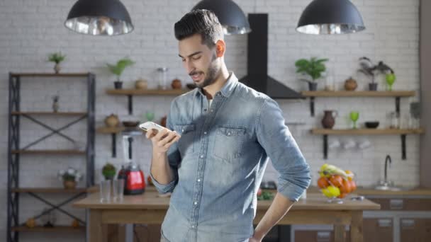 Portrait d'un jeune homme du Moyen-Orient qui enregistre et envoie des messages vocaux sur son smartphone à la maison. Heureux gars confiant parlant et souriant debout dans la cuisine à l'intérieur. Mode de vie. — Video