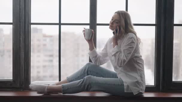 Ampla foto de mulher loira magro lindo sentado no peitoril da janela falando ao telefone e beber café. Retrato de relaxado despreocupado caucasiano millennial desfrutando manhã dentro de casa. — Vídeo de Stock