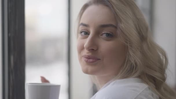 Retrato de close-up da bela mulher loira caucasiana com olhos grisalhos olhando para a câmera e pela janela. Feliz millennial despreocupado desfrutando de manhã em casa dentro de casa com xícara de café. — Vídeo de Stock