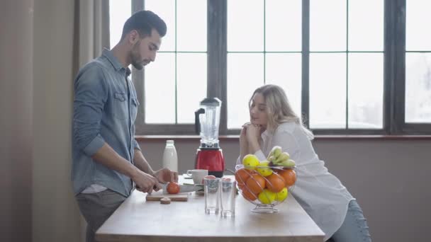 Feliz pareja interracial hablando y riendo cocinando ensalada saludable en casa. Retrato de vista lateral de la relajada mujer caucásica alegre y el hombre de Oriente Medio disfrutando de fin de semana juntos en el interior. Amor.. — Vídeo de stock