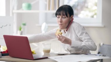 Öğle yemeği yiyen, telefonla konuşan ve ofiste dizüstü bilgisayarda klavye çalan genç bir kadın. İş yerindeki aşırı yüklenmiş beyaz tenli çalışanın portresi. Sağlıksız yaşam tarzı ve iş hayatı.