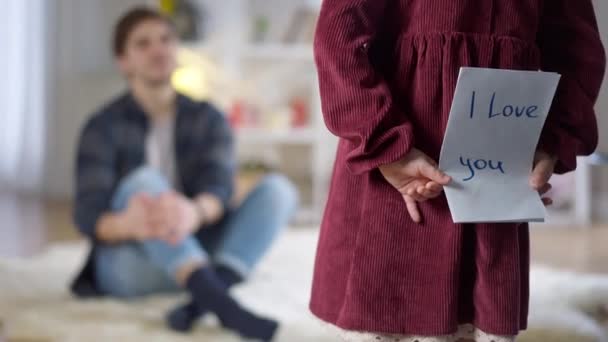 Back view of little Caucasian girl walking to smiling man giving card with I love you written. Happy loving daughter expressing feelings to single father at home indoors. Family concept. Stock Footage