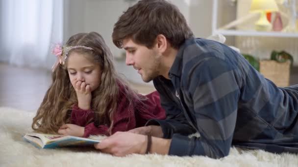 Linda chica escuchando al hombre leyendo el libro acostado en una alfombra suave en la habitación de la familia. Retrato de linda hija caucásica positiva disfrutando de la literatura con el padre en casa en el interior. Estilo de vida y hobby. — Vídeo de stock