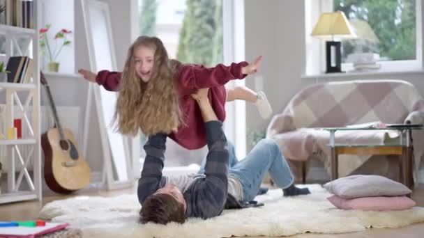 Emocionada niña juguetona imitando vuelo de avión como hombre feliz sosteniendo niño en las manos arriba. Retrato de la alegre hija caucásica jugando con el joven padre soltero en casa los fines de semana. — Vídeos de Stock