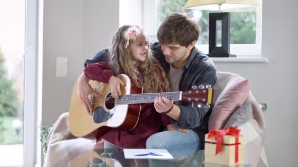 Ayah muda positif mengajar anak perempuan yang cukup lucu bermain gitar. Potret bahagia Kaukasia pria dan gadis duduk di sofa di rumah berbicara dan tersenyum. Konsep keibuan. — Stok Video