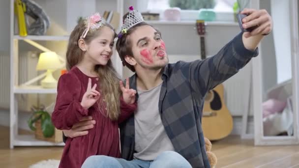 Veselý mladý muž s legrační make-up přičemž selfie s roztomilou dceruškou na smartphonu. Pozitivní kavkazský svobodný rodič baví s docela hravou dívku doma o víkendu. — Stock video
