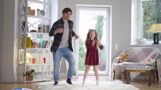 Funny dance of cheerful young father and playful cute daughter indoors at home. Wide shot of happy carefree Caucasian man and girl having fun together. Relaxation and family leisure. — Stock Video