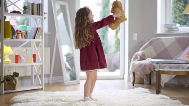 Ευρύ πλάνο πορτρέτο του χαρούμενου Καυκάσιου κοριτσιού με σγουρά μαλλιά που στριφογυρίζει με αρκουδάκι που αγκαλιάζει παιχνίδι. Σίγουρα χαμογελαστό χαριτωμένο παιδί απολαμβάνει την άνεση στο σπίτι σε εσωτερικούς χώρους. Τρόπος ζωής και παιδική ηλικία. — Αρχείο Βίντεο