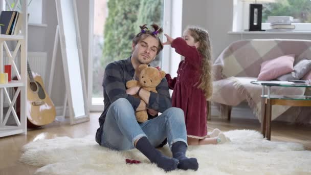 Portrait large de jeune homme patient assis sur un tapis mou dans le salon comme fille mignonne ludique faisant des queues de cheval pour le père. Brunette homme caucasien passer le week-end avec sa fille à la maison. — Video