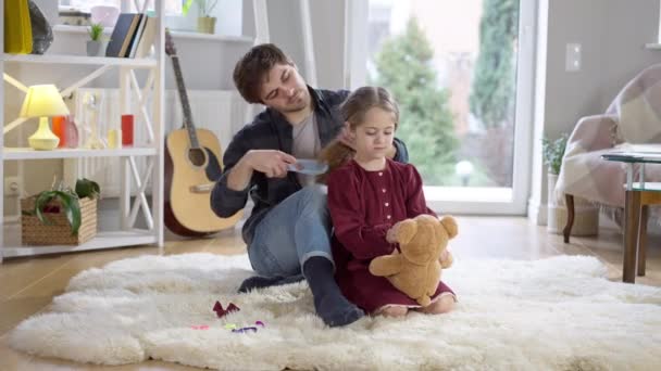 Amplio tiro de chica bonita feliz jugando con el oso de peluche como padre cariñoso peinando el pelo rizado largo de la hija. Morena Caucásico niño con padre y juguete en casa en interiores. Concepto de cuidado familiar. — Vídeos de Stock