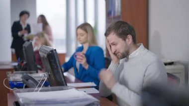 Bilgisayarın klavyesinde konuşan ve arka planda çalışan bulanık kadınlar olan konsantre beyaz adamın yan görüntüsü. Endişeli erkek çalışanın iş yerinde internetten mesajlaşması..