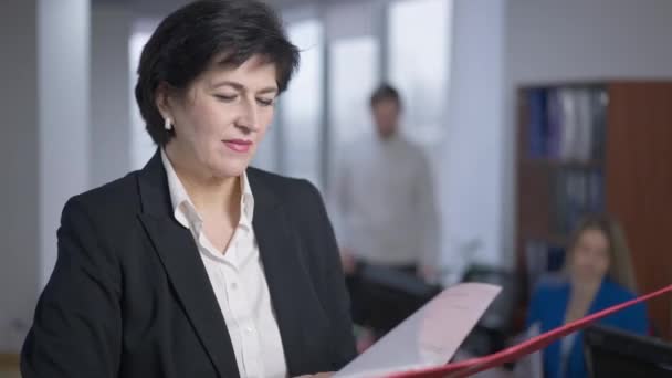 Retrato de mulher de negócios caucasiana concentrada elegante examinando documentos e olhando para a câmera sorrindo em pé no escritório com pessoas desfocadas no fundo. CEO confiante posando no local de trabalho. — Vídeo de Stock