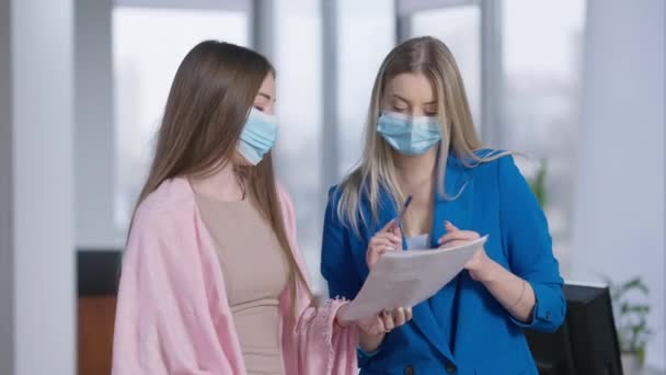 Dvě mladé krásné sebevědomé ženy v Covid face maskách diskutují o projektu v úřadu s rozmazaným mužem procházející v pozadí. Hubené blond a brunetky zaměstnanci mluví na koronavirus pandemie uvnitř. — Stock video