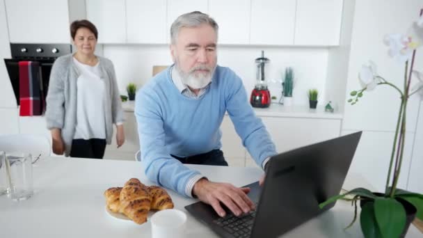 Portrait d'homme caucasien âgé occupé messagerie en ligne sur ordinateur portable comme femme aimante étreignant mari souriant et parlant. Heureux couple de retraités surfant sur Internet et bavardant dans la cuisine à la maison. — Video